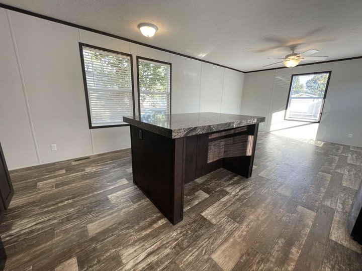 A modern, unfurnished room with wood-patterned flooring, white walls, large windows with blinds, and a dark wooden kitchen island under a ceiling light. A ceiling fan is visible in the adjoining space.