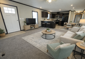 Modern open-concept living room and kitchen with neutral decor, light carpet, a beige sofa, armchair, round coffee table, TV on a stand, and a kitchen with dark cabinets and stainless steel appliances in the background.