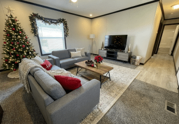 A cozy living room with gray sofas, a wooden coffee table, a TV on a stand, and a decorated Christmas tree by the window. Garland hangs above the window, and festive ornaments and a poinsettia adorn the space.