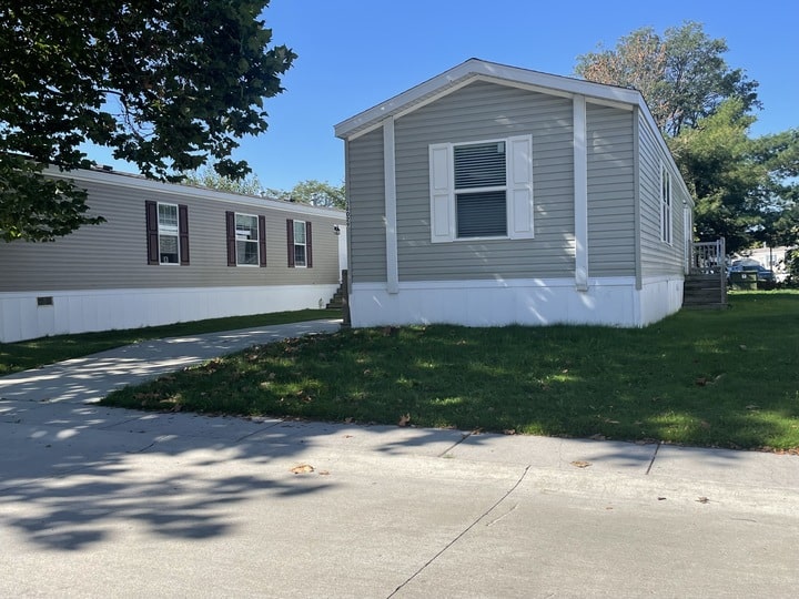 A gray manufactured home with white trim and shutters sits on a small grassy yard with a concrete driveway. Another similar home is adjacent, with trees and a clear blue sky in the background.