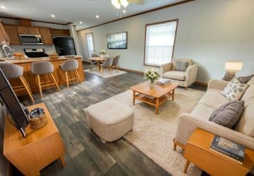 A modern open-concept living space with a cozy beige sofa, light wood furniture, a kitchen with bar stools, and a dining area. The room has wood flooring, bright windows, and neutral décor.