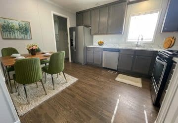 A modern kitchen with dark wood cabinets, stainless steel appliances, and a dining table set with green chairs on white rugs. Sunlight shines through a window above the sink, brightening the wood floor.