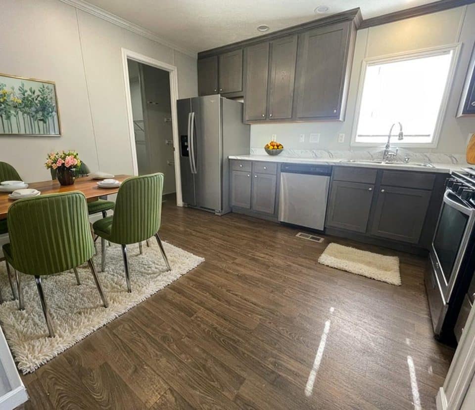 A modern kitchen with dark wood cabinets, stainless steel appliances, and a dining table set with green chairs on white rugs. Sunlight shines through a window above the sink, brightening the wood floor.