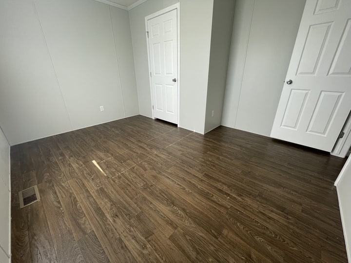 Empty room with light gray walls, dark wood laminate flooring, and two white doors; one door is closed and the other is partially open. A floor vent is visible in the lower left corner.