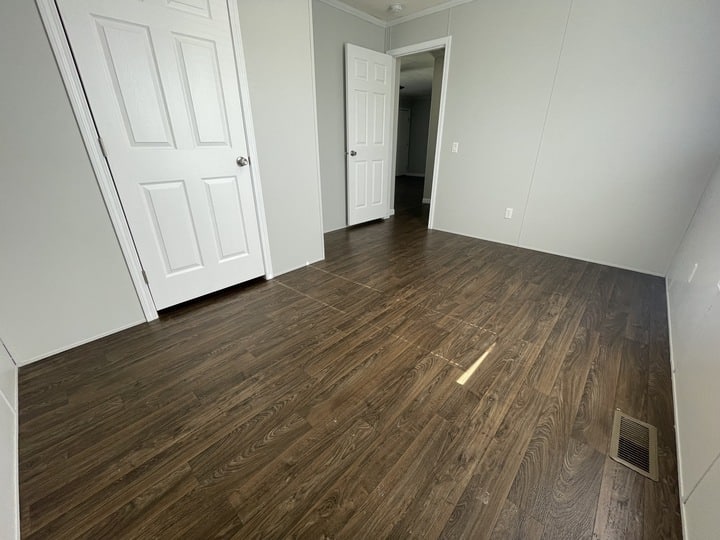 A small, empty room with white walls, two white doors, and dark wood flooring. One door is open, leading to another room with similar floors. A vent is visible on the floor near the wall.