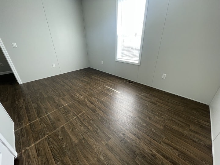 An empty room with light gray walls, a large window with blinds, and dark wood-style laminate flooring. Sunlight streams through the window, creating bright lines on the floor.