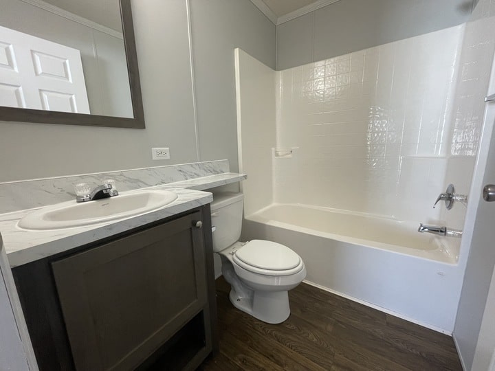 A small bathroom with a white bathtub and shower, a toilet, and a dark wood vanity with a sink and marble countertop. The floor is wood, and the walls are painted light gray. A mirror hangs above the sink.