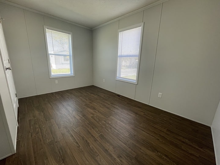 Empty room with gray walls, two large windows with blinds, and dark wood flooring. Sunlight enters through the windows, casting soft light on the floor. The room appears clean and simple.