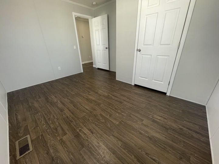 A small, empty room with light gray walls, dark wood flooring, and two white doors. An open doorway leads to a hallway. A floor vent is visible in the lower left corner.