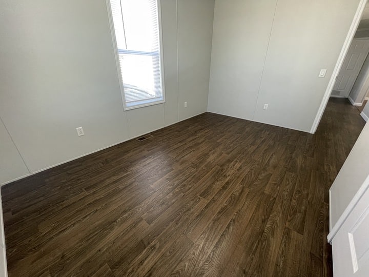 An empty room with light-colored walls, a single window with blinds, multiple electrical outlets, and dark wood flooring. The room is well-lit by natural light coming through the window.