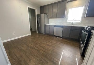 Kitchen with dark wood cabinets, stainless steel appliances including a fridge, dishwasher, and oven, marble countertops, and a window above the sink; wood-look flooring and light gray walls.