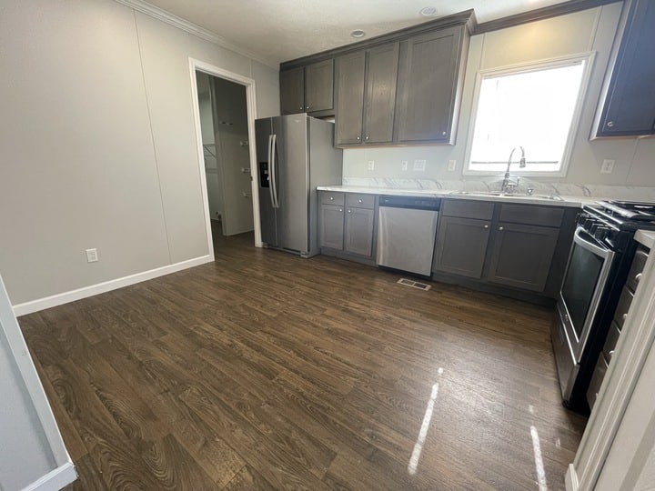 Kitchen with dark wood cabinets, stainless steel appliances including a fridge, dishwasher, and oven, marble countertops, and a window above the sink; wood-look flooring and light gray walls.