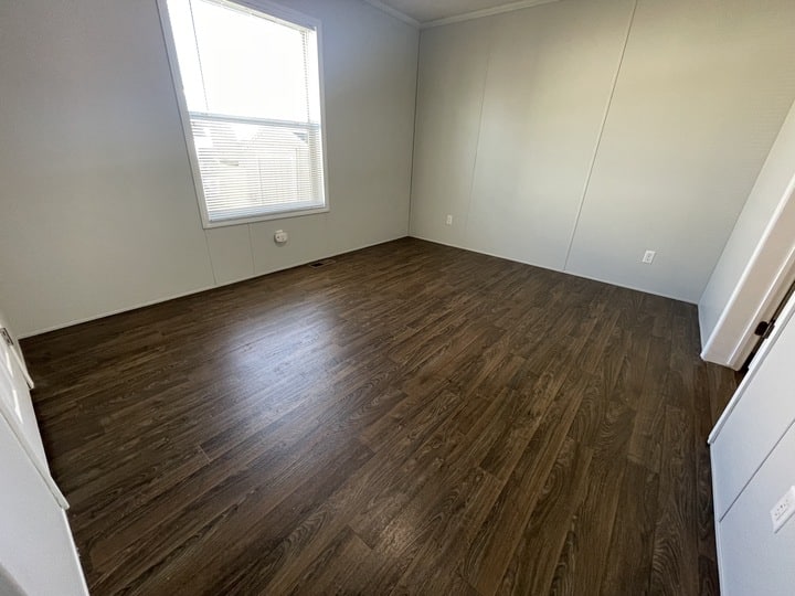 An empty room with dark wood flooring, light-colored walls, a large window letting in natural light, and white electrical outlets along the walls.