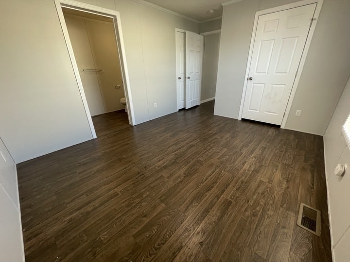 Empty room with dark wood flooring, light gray walls, white doors, a closet, and an open door leading to a small bathroom. The room appears clean and well-lit by natural light.