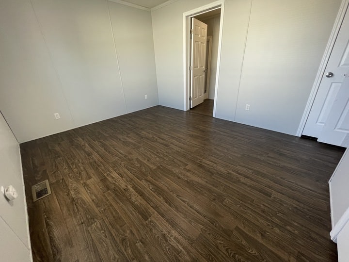 Empty room with dark wood flooring, light-colored walls, and white trim. Two white doors are visible, one open leading to another room and one closed. There is a floor vent on the left side.