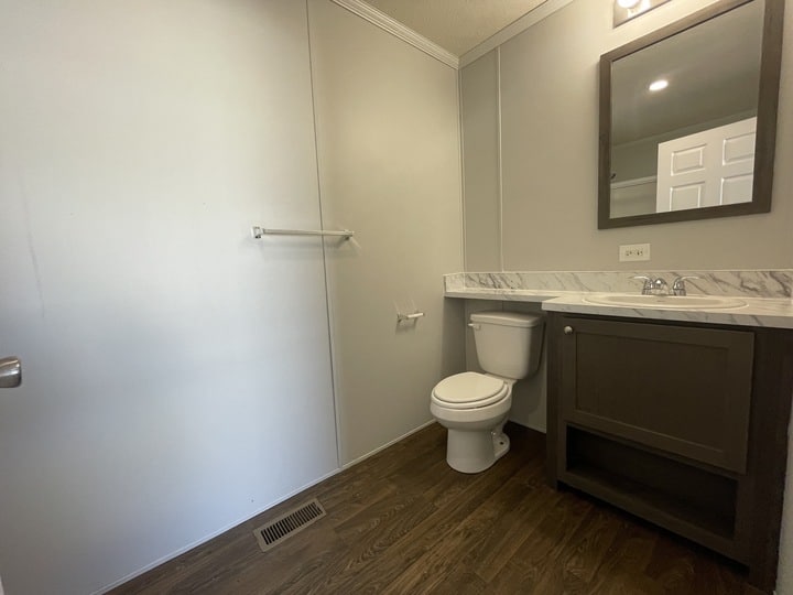 A simple bathroom with gray walls, wood-style flooring, a white toilet, a vanity with a sink and dark cabinet, a wall mirror, and a towel rack on the empty wall. The room has a clean and minimal design.