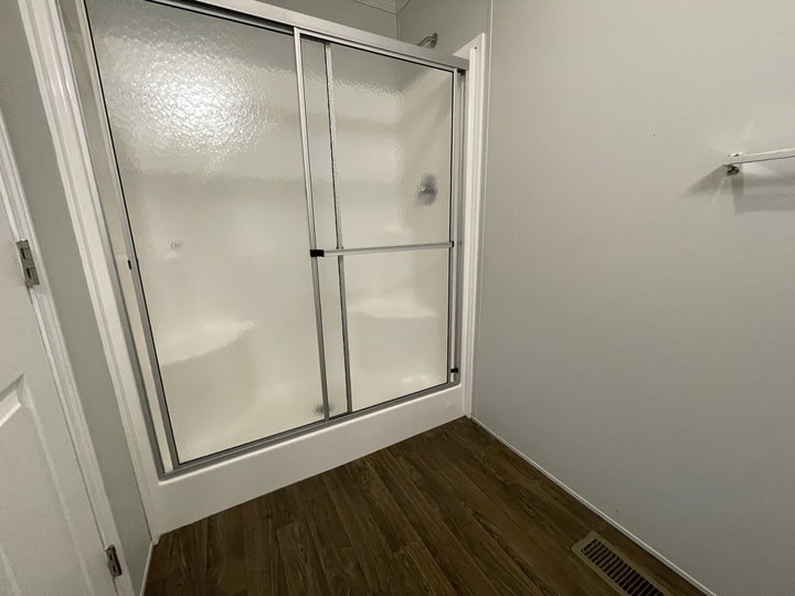 Bathroom with a glass-enclosed shower featuring sliding doors and built-in corner seats. The floor is dark wood, and the walls are light-colored. There is a white towel rack and a floor vent visible on the right side.