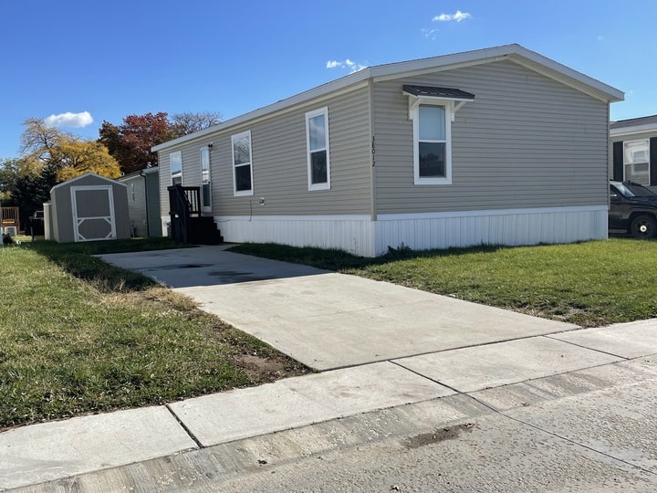 A beige manufactured home with white trim sits on a grassy lot, featuring a small front porch, a concrete driveway, and a matching storage shed in the background under a clear blue sky.