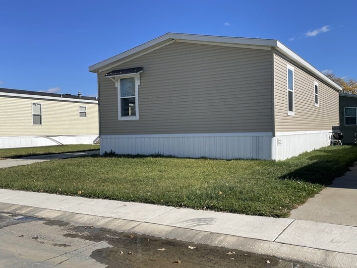 A beige manufactured home with white trim and a small window sits on a grassy lot next to a sidewalk, under a clear blue sky. Another similar home is visible in the background.