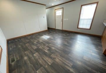 Empty room with gray wood-look vinyl flooring, light gray walls with brown trim, a ceiling fan, a window with blinds, and a white entry door. There is natural light coming through the window.