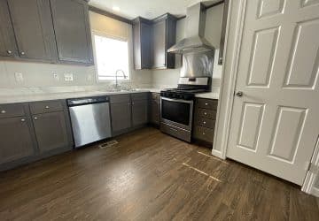 Modern kitchen with dark wood cabinets, stainless steel appliances including a dishwasher and oven, marble countertops, a vent hood, a window over the sink, and wood-look flooring. Door is closed on the right.