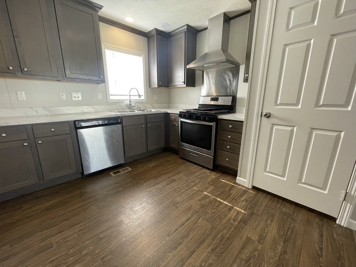 Modern kitchen with dark wood cabinets, stainless steel appliances including a dishwasher and oven, marble countertops, a vent hood, a window over the sink, and wood-look flooring. Door is closed on the right.