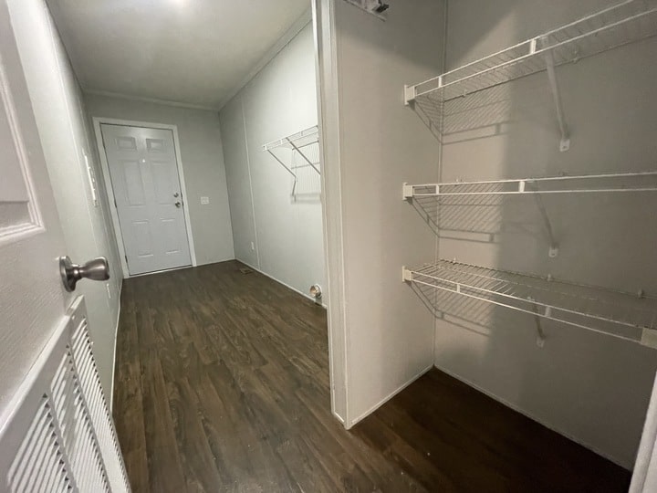 A narrow laundry or utility room with dark wood flooring, white walls, wire shelves on the right, and a white exterior door at the far end. The room is empty and brightly lit.