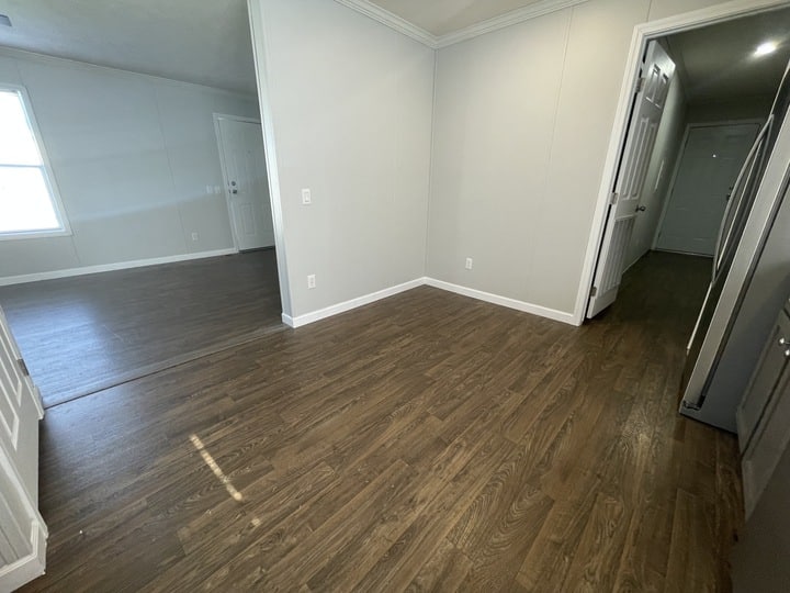 An empty room with light gray walls, white trim, and wood-style flooring. A doorway leads to a hallway and another space with a window is visible through an open entryway. The area appears clean and unfurnished.