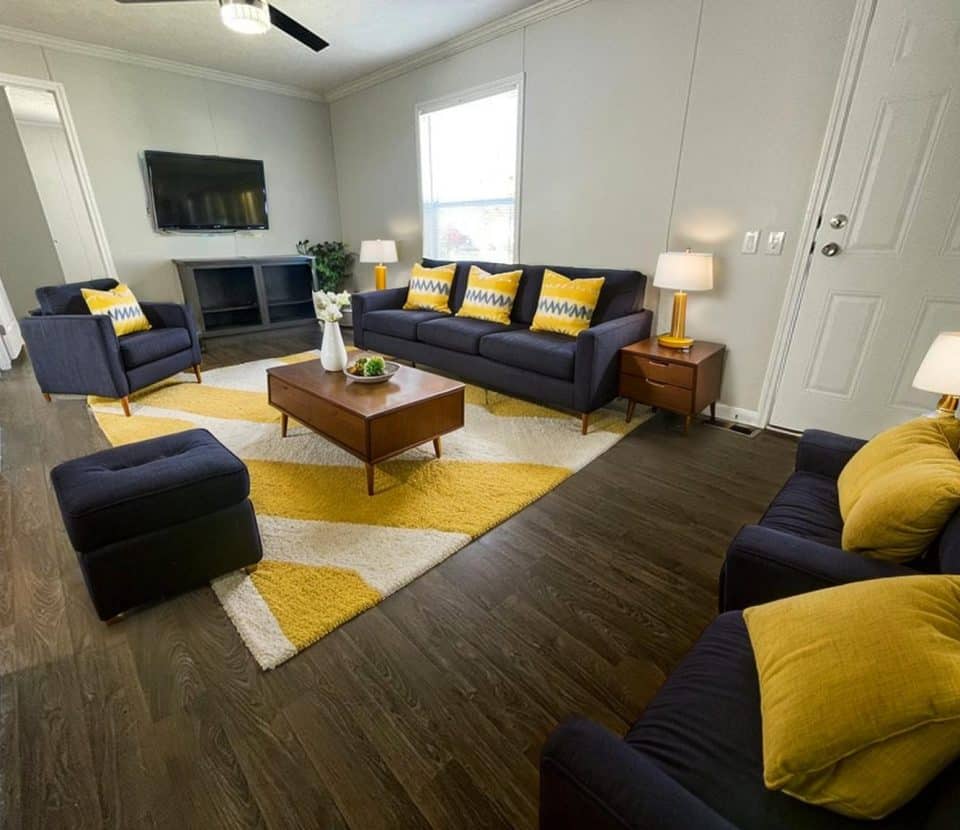 A modern living room with dark blue sofas, yellow and white accent pillows, a wooden coffee table, yellow and white geometric rug, three table lamps, wall-mounted TV, and a window letting in natural light.