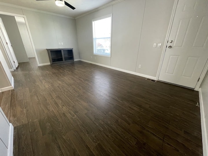 Empty room with dark wood flooring, light gray walls, a white door, a window letting in natural light, a ceiling fan, and a small built-in shelf unit against one wall.