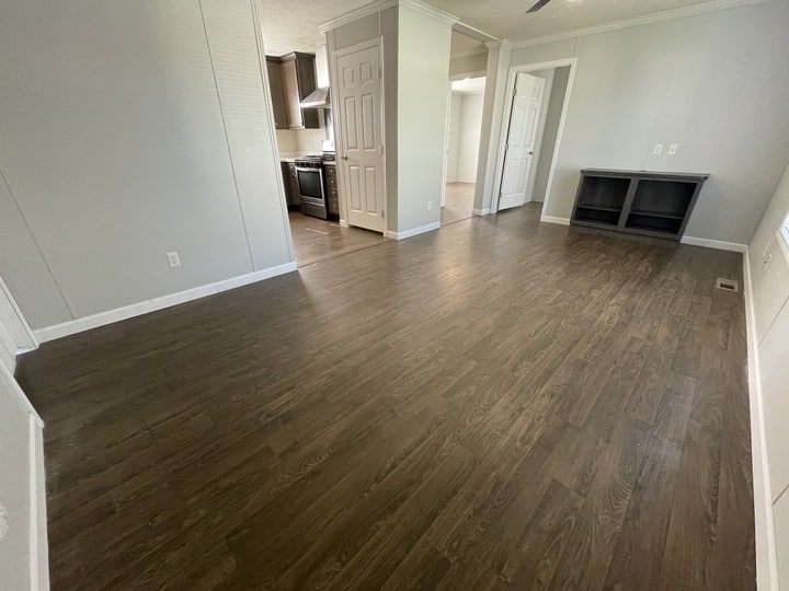 A spacious, empty room with dark wood flooring, white walls, and doorways leading to a kitchen and other rooms. There is built-in shelving on one wall and natural light coming from an unseen window.