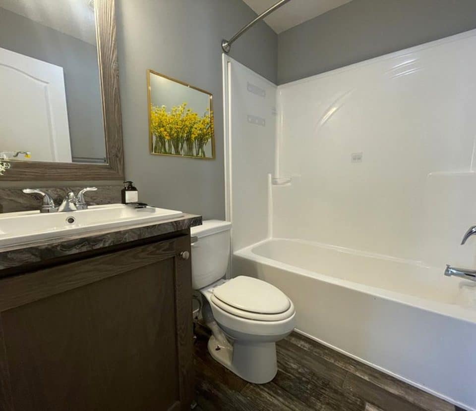 A modern bathroom with a white bathtub and shower, gray walls, a toilet, a sink with a dark wood vanity, a large mirror, and a wall art featuring yellow flowers above the toilet.