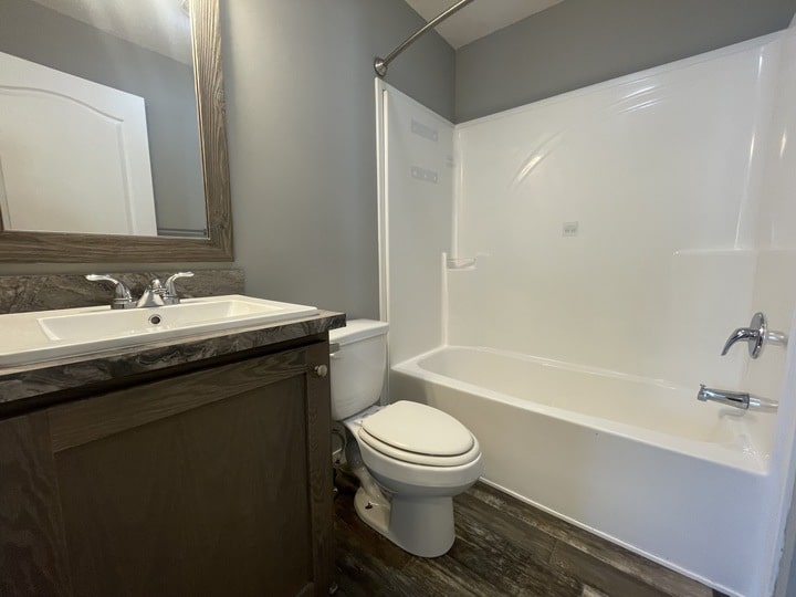 Modern bathroom with gray walls, a white bathtub-shower combo, a toilet, and a dark wood vanity with a white sink and chrome faucet. A large mirror hangs above the sink, and the floor has a dark tile pattern.
