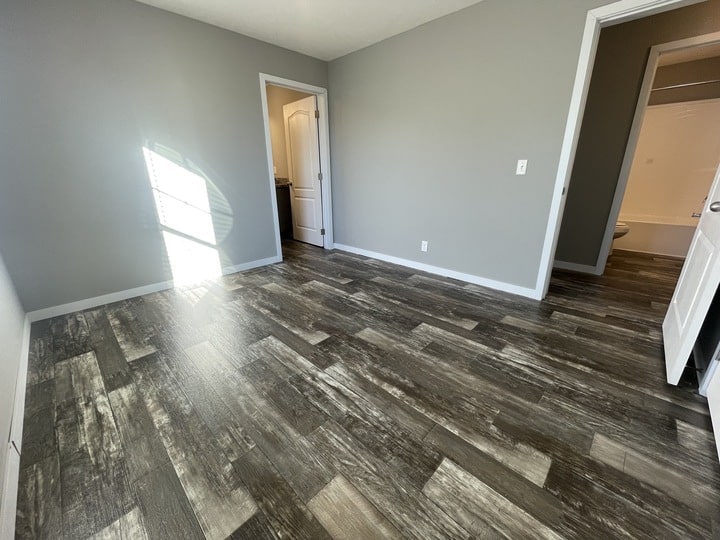 A small, empty room with gray walls, dark wood-patterned flooring, and two open doors—one leading to a bathroom and another to a hallway. Sunlight streams in through a window, casting shadows on the floor.