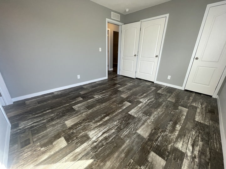 Empty room with gray walls, dark wood-patterned flooring, white baseboards, and two closed white doors. Sunlight shines across the floor from a window out of view.