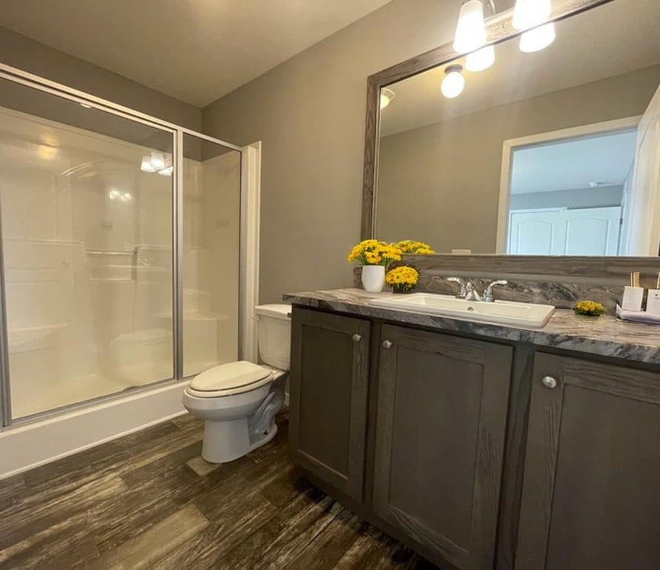 Modern bathroom with dark wood cabinets, a large mirror, a sink, toilet, and a glass-enclosed shower. The countertop has yellow flowers and toiletries. The room has neutral walls and wood-patterned flooring.