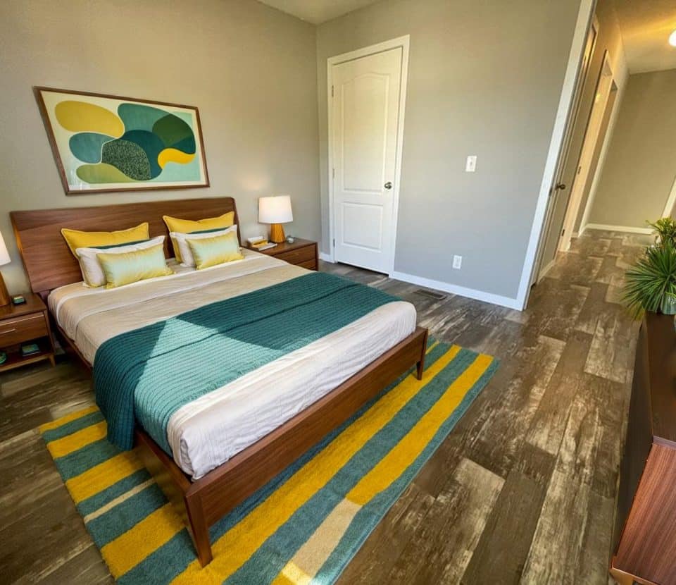 A modern bedroom with a wooden bed, teal and yellow bedding, two bedside tables with lamps, a colorful abstract painting above the bed, a striped rug, and a dresser with plants and books. Brown floor, light gray walls.