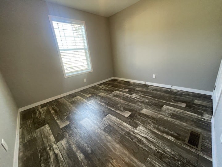 An empty room with gray walls, a single window with white blinds, and dark wood-style flooring. There are white baseboards and electrical outlets along the walls.