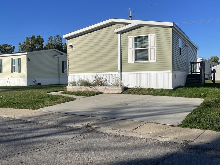 A light green manufactured home with white trim and shutters sits on a grassy lot. A concrete driveway and small flower bed are in front, with another similar home visible to the left. The sky is clear and blue.