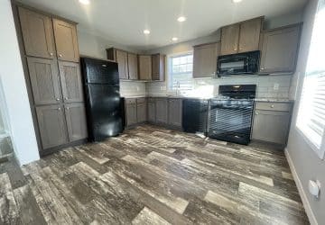 Modern kitchen with dark wood cabinets, black appliances (fridge, dishwasher, oven, microwave), light tile backsplash, wood-look flooring, and a window letting in natural light. Room is clean and spacious.