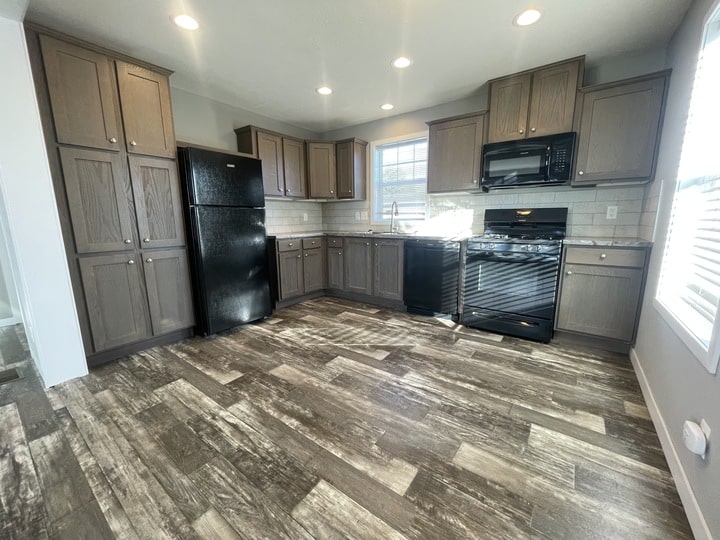 Modern kitchen with dark wood cabinets, black appliances (fridge, dishwasher, oven, microwave), light tile backsplash, wood-look flooring, and a window letting in natural light. Room is clean and spacious.