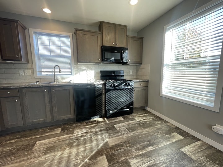 Modern kitchen with wood cabinets, a black stove and microwave, dishwasher, tiled backsplash, and large windows letting in natural light, with wood-look flooring.