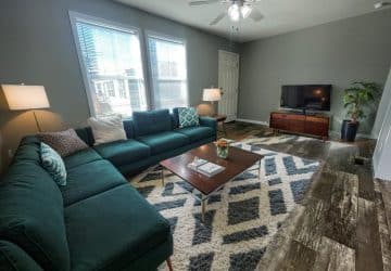 A modern living room with a green sectional sofa, geometric rug, wooden coffee table, two lamps, TV on a wooden stand, a plant, and large windows letting in natural light. The walls are gray and the floor is wood.