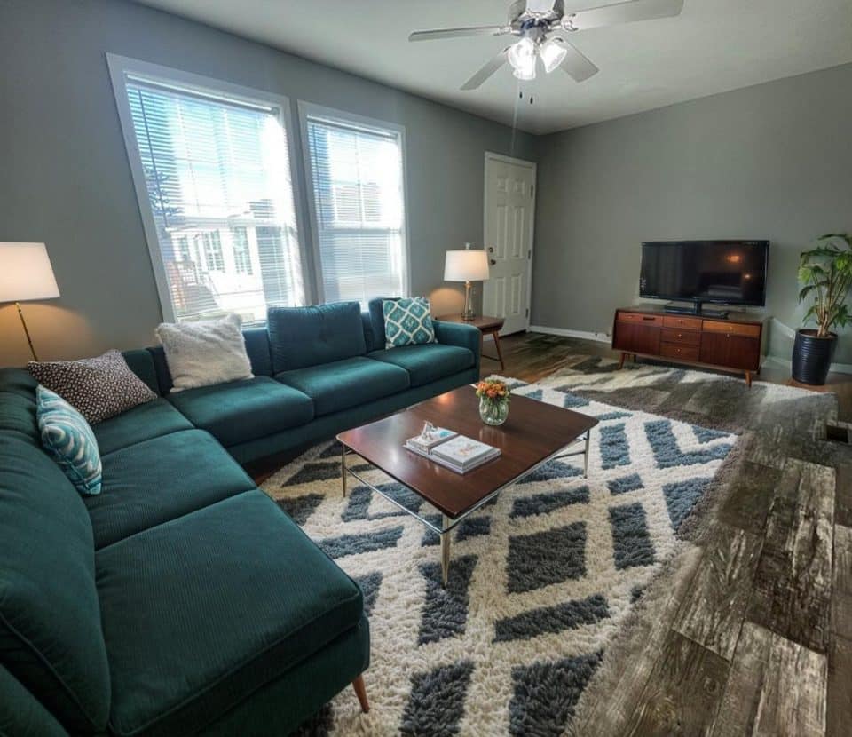 A modern living room with a green sectional sofa, geometric rug, wooden coffee table, two lamps, TV on a wooden stand, a plant, and large windows letting in natural light. The walls are gray and the floor is wood.