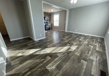Empty living room with gray wood-style flooring, light gray walls, and a white ceiling fan. An open doorway leads to a kitchen with stainless steel appliances and large windows letting in natural light.