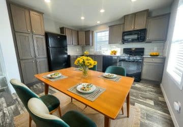 Modern kitchen with dark wood cabinets, black appliances, and a wooden dining table set for three. A vase of yellow flowers is centered on the table, and sunlight streams in through a window on the right.
