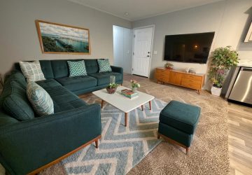 A modern living room with a green sectional sofa, patterned pillows, a white coffee table with books, a TV on a wooden console, plants, and a large wall photo above the sofa. The room has a gray carpet and light walls.
