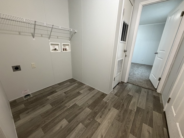 A small, empty laundry room with gray wood-look flooring, white walls, a wire shelf, washer and dryer hookups, and a door leading to a carpeted hallway.