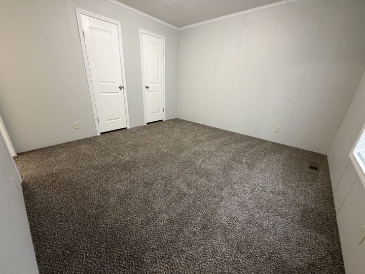 Empty room with gray carpet, white walls, two closed white doors, and a window letting in natural light from the right side. The space appears clean and unfurnished.