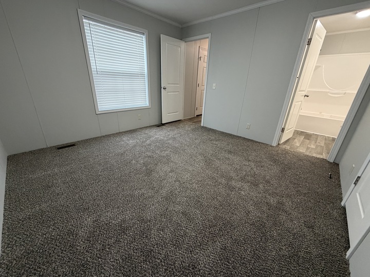 A bright, empty room with gray carpet, light gray walls, a large window with closed blinds, and two open white doors—one leading to a bathroom and the other to a closet or hallway.